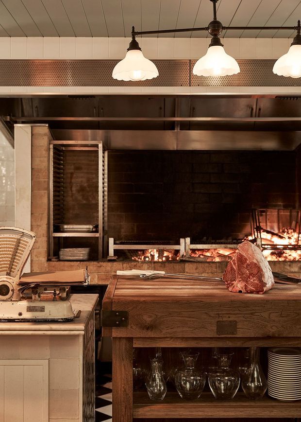 A warmly lit commercial-style cooking space featuring a large open wood-fired hearth, stainless-steel ventilation and shelving, vintage pendant lights, tiled walls, and a substantial dark timber butcher-block worktable. A meat slicer, glassware, stacked plates, and cooking equipment reinforce the restaurant kitchen setting.