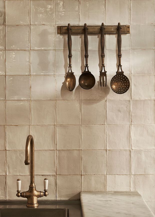 Close-up of a warm, rustic kitchen corner featuring a handmade cream tile splashback, an aged brass gooseneck tap, a dark sink, a marble benchtop, and vintage bronze-toned cooking utensils hanging from a wall rail.