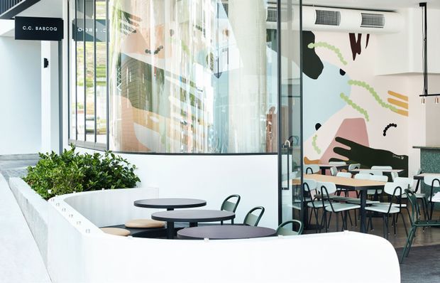 Bright contemporary commercial café or dining space viewed from an adjoining circulation area. The scene features curved white built-in planter and bench forms, black round tables, green metal chairs, extensive internal glazing, pendant lighting, and a large colourful abstract mural. A visible sign reads “C.C. BABCO” near the entrance.