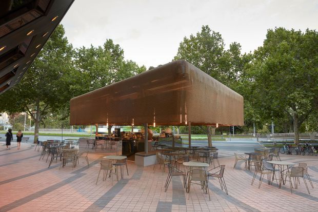 Exterior view of a contemporary pavilion or café structure in a landscaped urban plaza. The compact elevated building has a rounded rectangular form wrapped in warm copper-toned perforated metal screening, with open glazing and a service area beneath. Numerous lightweight outdoor tables and chairs occupy the surrounding paved terrace, with mature trees and a sports or public recreation setting in the background.