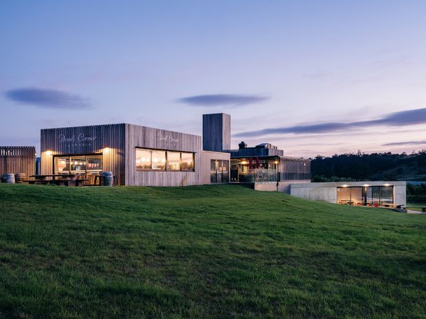A contemporary low-rise building complex set into a gently sloping grassy landscape at dusk. The architecture features several interconnected rectangular volumes clad in vertically ribbed grey metal, large illuminated glazed openings, a taller narrow tower element, and a partially recessed lower-level glazed volume. Outdoor seating is visible near the entrance, with dark trees in the background and a blue-purple evening sky.
