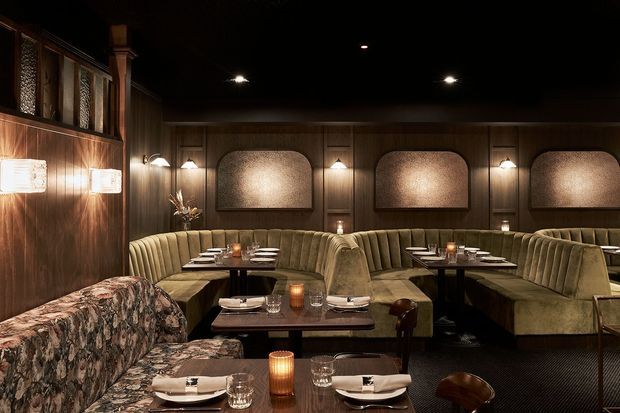 A dimly lit, richly detailed restaurant dining room with rows of curved upholstered banquette seating, dark timber wall panelling, framed textured feature panels, intimate table lighting, and formally set dark dining tables. The space has a moody, luxurious hospitality interior with a strong mid-century-inspired atmosphere.