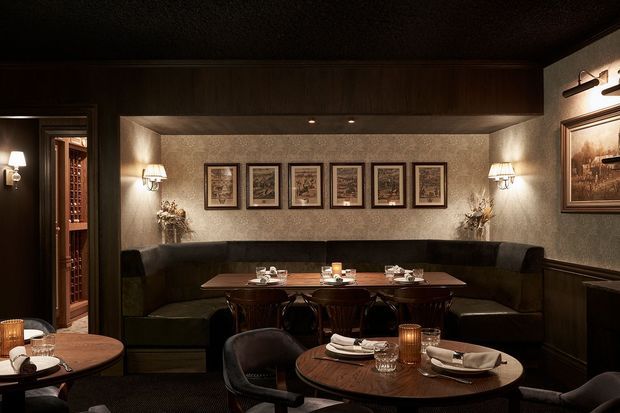 A dimly lit, intimate restaurant dining room with dark wood wall panelling, a large U-shaped upholstered banquette, round and rectangular timber dining tables, and upholstered dining chairs. Warm wall sconces and table lamps illuminate textured plaster walls decorated with framed artwork, creating a moody, refined hospitality interior.