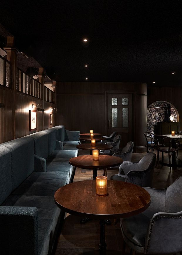 A dimly lit, atmospheric hospitality lounge or bar interior with dark timber wall panelling, a black textured ceiling, warm candlelit tables, a long upholstered banquette, and dark velvet armchairs. Round timber tables are arranged through the narrow seating area, with framed wall art, decorative lighting, and a glazed timber door adding character.