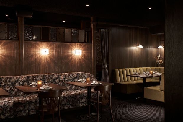 Moody, intimate restaurant dining area with dark timber wall panelling, decorative patterned upper screens, upholstered banquette seating, small set dining tables, and warm wall sconces. The space has a refined hospitality interior with rich brown tones and low ambient lighting.