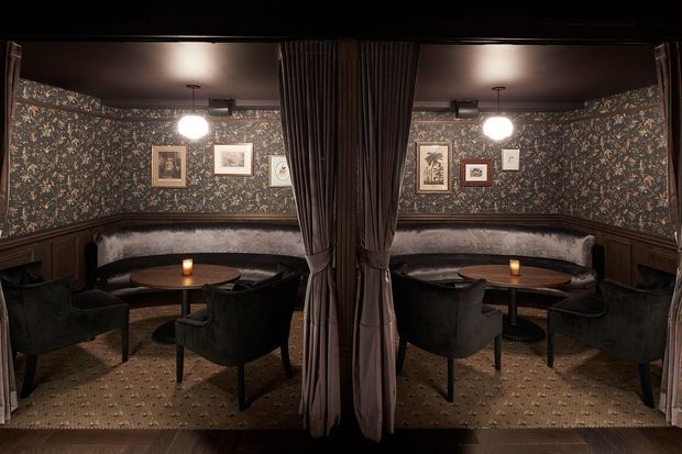 A dark, richly decorated lounge or hospitality space viewed symmetrically through a central curtain partition. The room features patterned botanical wallpaper, dark timber wainscoting, upholstered banquette seating, velvet armchairs, small round timber tables with candles, framed artwork, pendant globe lights, and a patterned carpet. A large mirror or reflective partition creates a mirrored appearance and reinforces the symmetry.