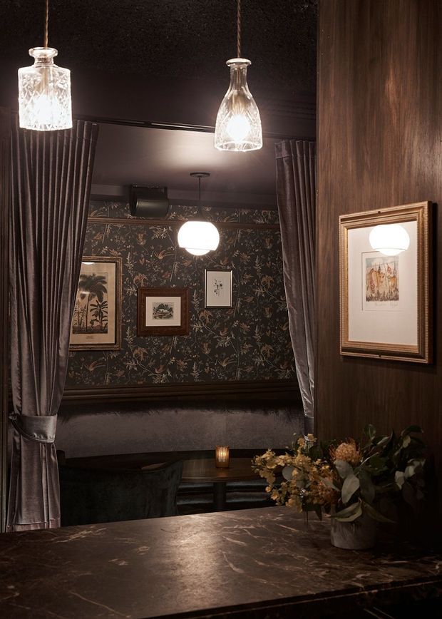 Moody, richly layered interior view through gathered curtains into a refined lounge or dining setting. The space features dark timber wall panelling, botanical patterned wallpaper, framed artwork, velvet upholstery, marble-like dark stone surfaces, and warm pendant and table lighting. The overall atmosphere is intimate, dramatic, and luxurious.