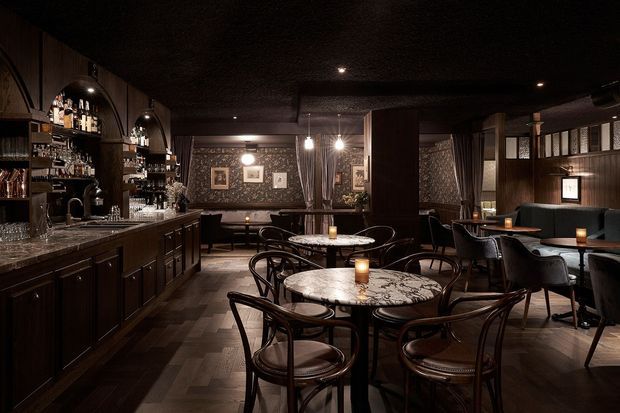 A moody, sophisticated hospitality interior featuring a dark timber bar with arched bottle shelving, marble countertops, round marble café tables, bentwood chairs, upholstered booth seating, patterned wall finishes, framed artwork, and warm pendant and candle lighting.