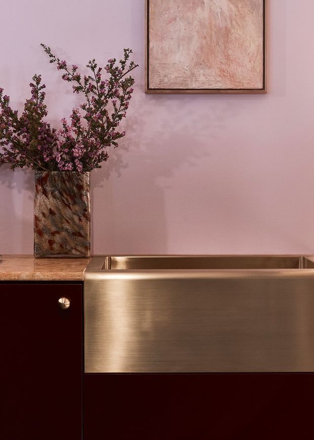 Close-up interior vignette featuring a brushed bronze or copper-toned integrated sink and cabinetry, with dark burgundy joinery, a warm pink plaster wall, a textured ceramic vase with flowering branches, and framed abstract artwork. The composition has a refined, moody, jewel-toned character.