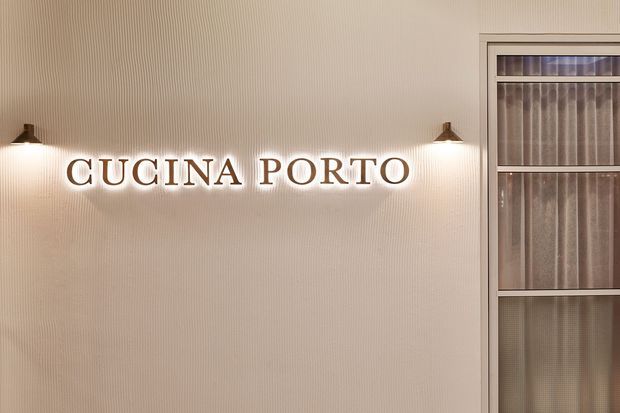 A close-up interior view of a softly lit wall featuring illuminated “CUCINA PORTO” signage in dark lettering, flanked by small wall sconces. The wall has fine vertical ribbed detailing, and a framed glazed partition or door with translucent textured panels is visible on the right. The overall presentation suggests a refined hospitality or restaurant interior.