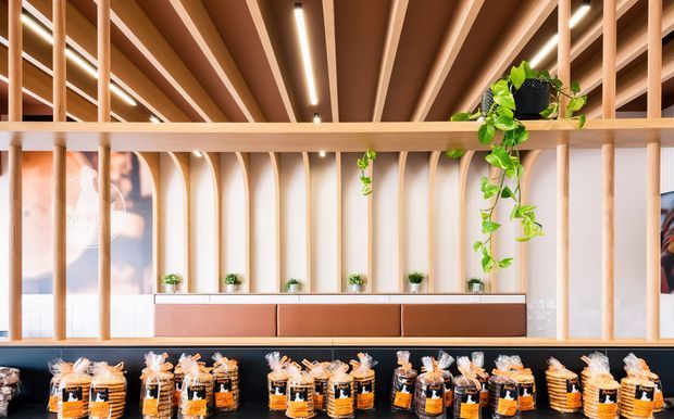 Bright contemporary commercial reception or retail display area featuring a long timber-toned counter, rows of packaged products, vertical timber screening, curved white wall fins, a slatted timber ceiling with integrated linear lighting, and trailing indoor greenery.
