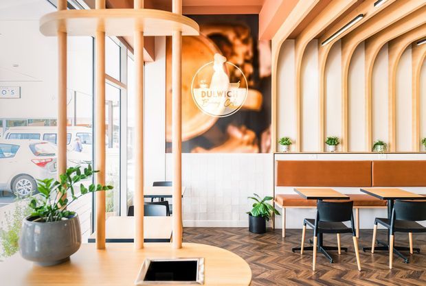 Bright contemporary café or hospitality interior with a warm timber fit-out. The space features a curved timber service or communal counter in the foreground, vertical timber columns, a tiled wall, built-in upholstered bench seating, small potted plants, black dining chairs, and herringbone timber flooring. Full-height glazing opens the space to the street, while a large Dulwich-branded wall graphic forms a focal point.