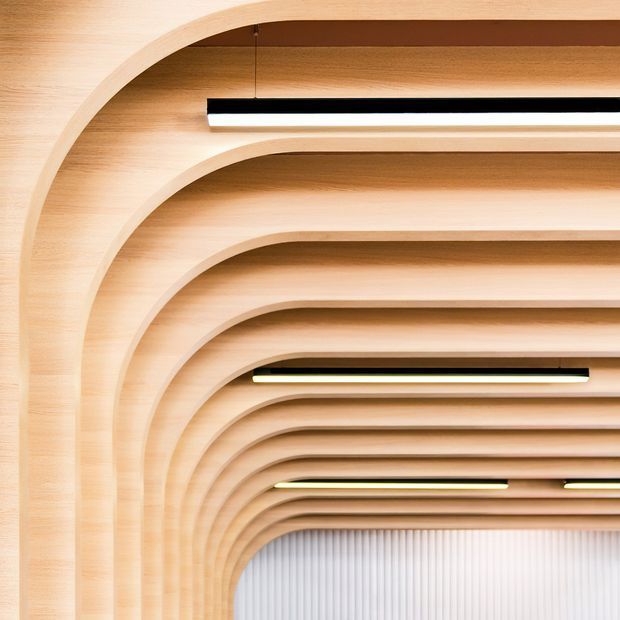 Close-up architectural view of a richly detailed timber-lined ceiling and wall transition, formed by repeated curved ribs or arches that create a tunnel-like perspective. Slim linear pendant lights are suspended between the ribs, and a vertically ribbed pale wall is visible at the far end. The image emphasizes warm wood craftsmanship, rhythm, and sculptural interior detailing.