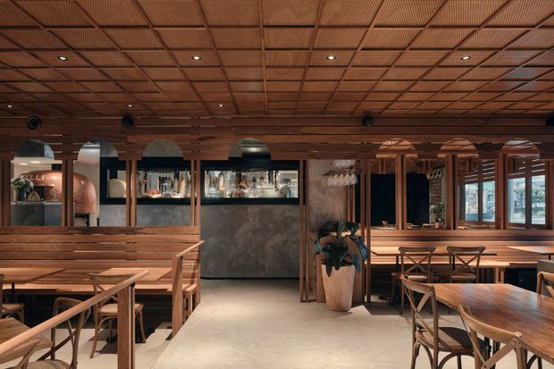 A warmly lit restaurant interior with extensive dark timber detailing, a gridded timber ceiling, built-in bar and display shelving, timber dining tables and chairs, and large windows along one side. The space has a refined rustic and contemporary hospitality character, with a central service counter and planted feature.