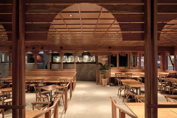 A warmly lit restaurant interior with extensive timber columns, beams, lattice ceiling detailing, and timber dining tables and chairs. The open dining area surrounds a central bar or service counter, with pendant and recessed lighting, woven cane-style seating, and polished concrete or stone-look flooring. The space has a refined tropical or resort-inspired character with strong natural timber detailing.