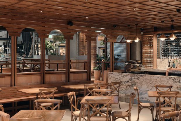 A warmly lit restaurant or hospitality interior with extensive timber wall panelling and a decorative timber coffered ceiling. The space includes banquette seating, wooden cross-back chairs, marble-clad bar counters, pendant lighting, mirrored panels, and a visible wine display behind the bar. The overall atmosphere is refined, warm, and inviting, with a strong contemporary rustic character.