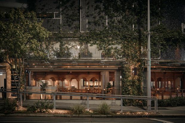 Street-facing view of a warmly lit commercial hospitality venue, likely a restaurant or café, with extensive timber-framed glazing, timber doors, a covered frontage, planted greenery, and an accessible ramp with metal handrails. The building frontage is integrated with climbing vegetation and illuminated landscaping, creating an inviting evening streetscape.