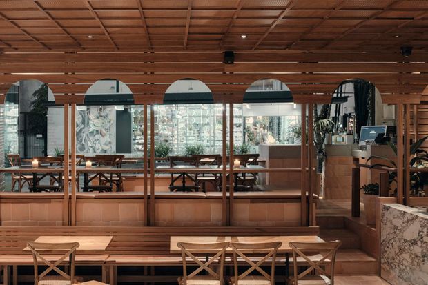 A warm, refined restaurant or hospitality interior viewed across a dining area. The space features extensive timber wall and ceiling panelling with a gridded exposed-structure effect, large arched-top windows, timber-framed glazing, built-in timber bench seating, small wooden dining tables, and cross-back chairs. A stone-clad service or bar counter occupies the right side, with additional dining and planting visible beyond the glazing.