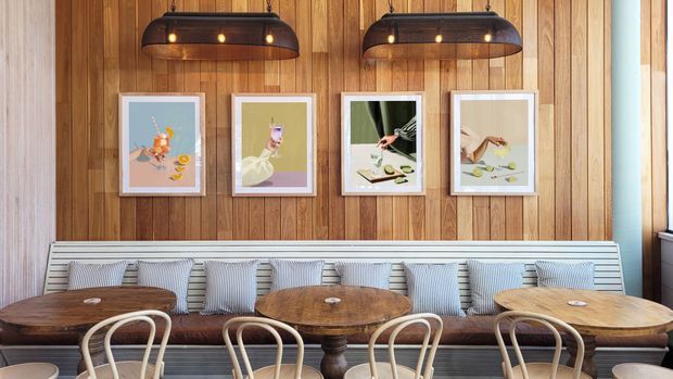 A warmly lit dining area with a long built-in upholstered banquette and multiple round wooden dining tables. The room features a full-height vertical timber feature wall, framed artwork, pendant lights with dark metal shades, striped cushions, and bentwood dining chairs.
