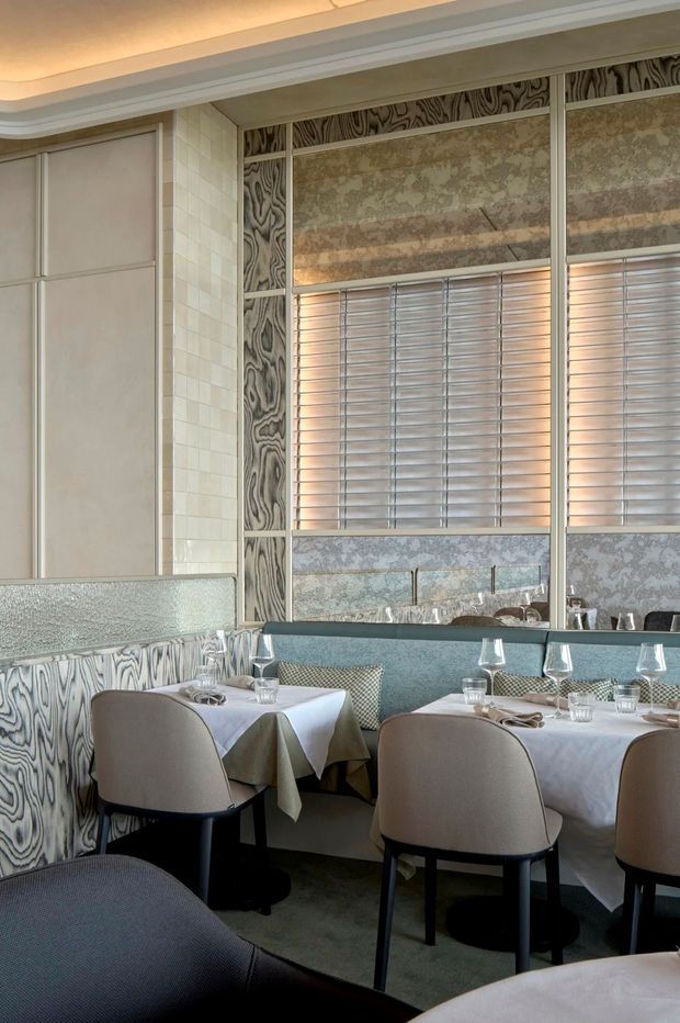 A refined restaurant dining room with closely arranged tables dressed in white linens, upholstered curved-back chairs, and a long upholstered banquette. The space features an ornate patterned wall treatment, pale tiled wall panels, large mirrored or glazed panels with horizontal blinds, and a layered recessed ceiling detail. The palette is muted beige, grey, cream, and pale blue-green, creating an elegant, softly lit atmosphere.