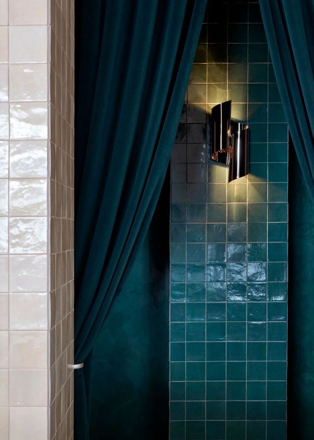 A close-up architectural detail of a moody bathroom or shower enclosure. The space features glossy teal square wall tiles, a contrasting vertical strip of pale white tiles, deep teal fabric curtains, and a sculptural black-and-chrome wall light with warm illumination. The composition is narrow and intimate, with rich jewel tones and reflective surfaces.