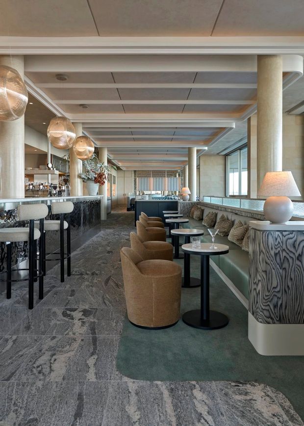 A refined hospitality lounge and bar interior with a long banquette, small round café tables, upholstered tan chairs, marble-look flooring, and a dark stone bar. Large cylindrical columns, extensive windows, layered ceiling beams, woven pendant lights, table lamps, and patterned decorative joinery create a luxurious contemporary setting.
