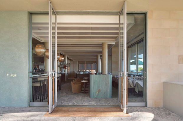 View through open glazed doors into a refined restaurant or hospitality dining space. The entrance has pale stone or rendered walls, slim metal-framed glazing, and a timber threshold. Inside, the long room features exposed timber ceiling slats and beams, columns, pendant lighting, upholstered dining chairs, tables, a central blue-green host counter, and patterned stone or tile flooring. The image is taken from outside, with the building entrance and interior dining area visible.