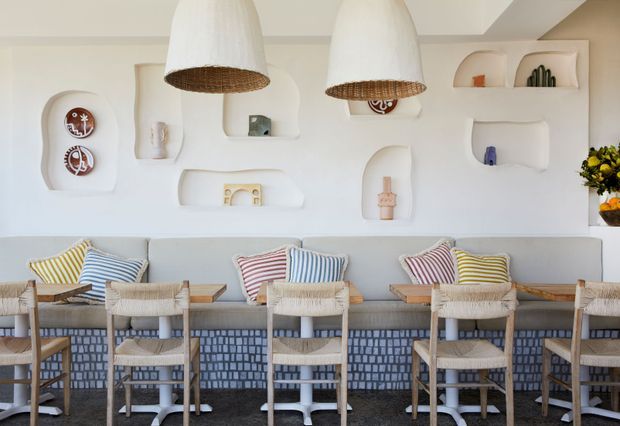 A bright, eclectic café or hospitality interior with a long built-in counter and bar seating. The white sculptural plaster wall features multiple rounded recessed display niches containing small decorative objects. Large woven pendant lights hang above the counter, while pale timber and woven bar stools pair with colourful striped cushions. The counter front has a distinctive blue-and-white patterned tile finish, creating a playful coastal Mediterranean atmosphere.
