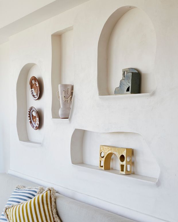 Bright, softly styled interior wall with multiple arched recessed display niches containing small decorative objects and ceramics. A pale sofa with striped cushions is visible along the bottom edge. The wall has a sculptural, plastered finish and a warm minimalist decorative character.