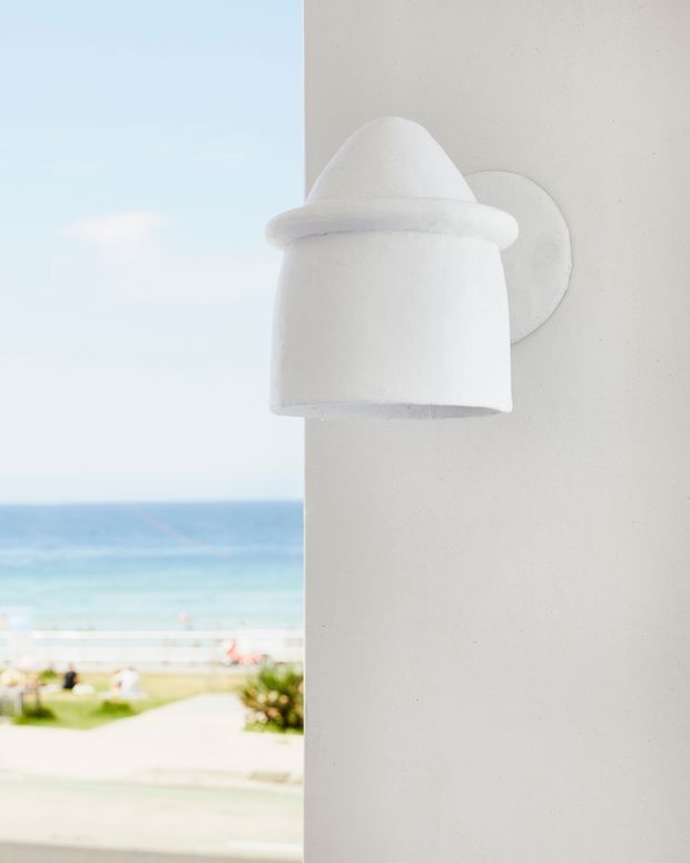 Close-up of a white sculptural wall-mounted light fixture on a smooth white exterior wall, with a softly blurred beach, ocean, promenade and landscaped grounds visible beyond the building edge. The image has a bright, minimal coastal atmosphere.