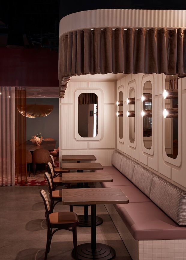 A refined, moody restaurant dining interior with a long upholstered banquette, small wood-topped tables, dark timber-framed chairs, and warm decorative lighting. The room features cream fluted wall panels, rounded architectural surrounds, repeated inset mirrors, and a dramatic curved ceiling canopy trimmed with dark pleated fabric. Deep brown, blush, mauve, and cream tones create an intimate, luxurious atmosphere.
