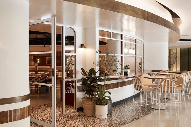A refined open-plan hospitality or commercial interior featuring a curved white service counter and curved ceiling bulkhead, framed glass partitions, terrazzo flooring, timber detailing, potted plants, and a light dining area with café tables and chairs. A warmer dining or lounge space is visible through the glazed partition.