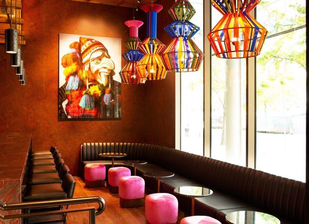 A richly coloured contemporary hospitality interior, likely a bar or restaurant lounge, with a long curved upholstered banquette, small round tables, black leather seating, vivid pink stools, timber flooring, dark reddish-brown walls, and sculptural multicoloured pendant lights. Full-height glazing brings in strong daylight, while a colourful artwork adds a bold decorative focal point.
