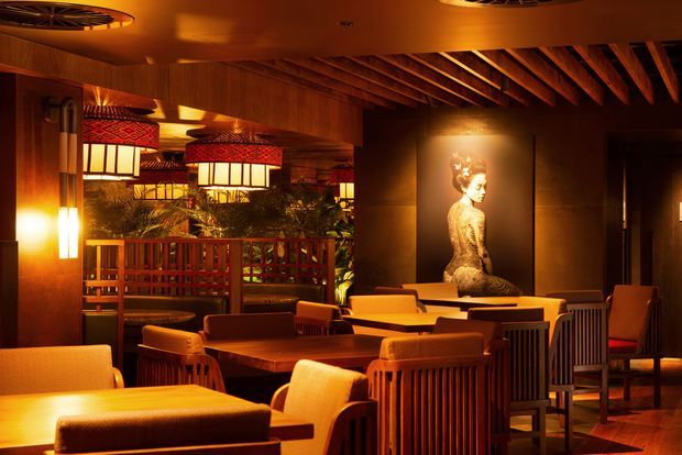 A warmly lit, richly detailed restaurant or hospitality interior with timber wall panelling, exposed timber ceiling beams, decorative pendant lanterns, timber dining tables and chairs, and a dramatic portrait feature wall. The space has a sophisticated, intimate atmosphere with extensive warm amber lighting and planted screening around a raised seating area.