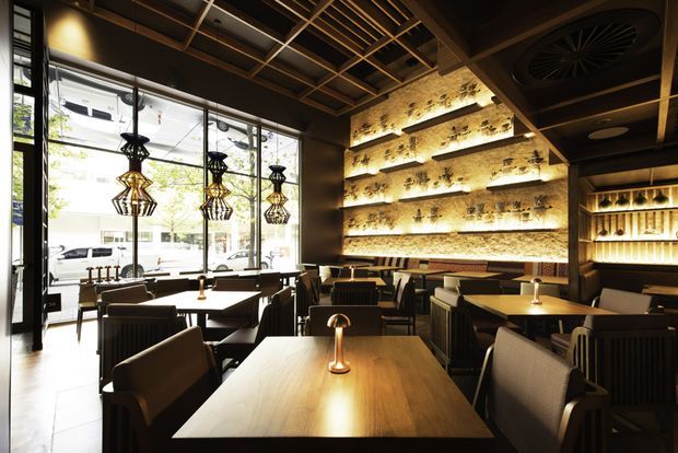 A warmly lit contemporary restaurant or hospitality interior with rows of dark upholstered dining chairs and timber tables. The space features large street-facing windows, decorative pendant lights, a dramatic backlit textured stone feature wall with shelving and plants, illuminated bottle displays, and a dark open-grid ceiling with exposed services.