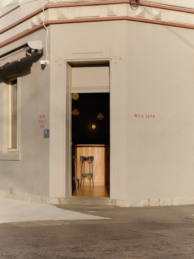 Street-level exterior view of a heritage-style corner commercial building with a recessed entrance to the Mona Public Bar. The pale rendered facade has decorative horizontal banding, raised geometric detailing, timber-framed openings, red painted lettering, and a small glimpse of the bar interior with timber joinery and pendant lights.