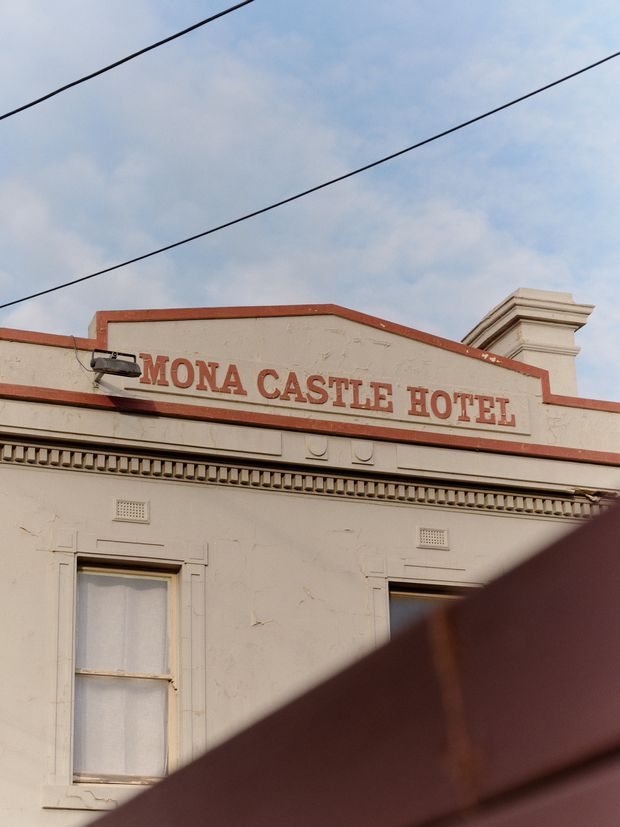 Street-level close-up of the Mona Castle Hotel facade, showing a cream rendered exterior with red-painted trim, raised red lettering, decorative cornice and balustrade-like detailing, tall sash windows, and a prominent chimney. Overhead utility wires cross the sky, while a dark foreground obstruction partially covers the lower right portion of the building.