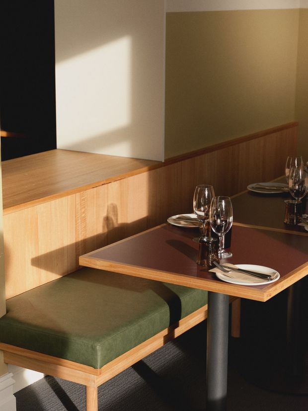 A warmly lit restaurant dining booth with green upholstered bench seating, timber wall panelling and built-in timber ledges. Dark rectangular dining tables are set with plates, cutlery and wine glasses, while strong sunlight creates geometric shadows across the wood and seating. The space has a refined, intimate hospitality interior.