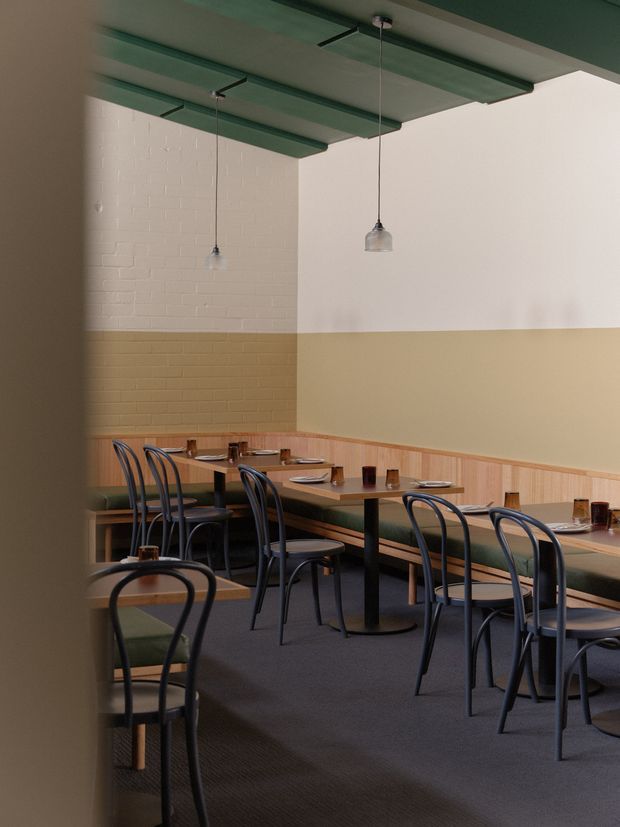 A refined restaurant dining area with rows of dark bentwood-style chairs and square tables set with plates and glasses. Built-in banquette seating lines the walls, which feature a two-tone white and ochre painted brick finish with timber wall panelling below. The dark green ceiling has exposed beams or panels and suspended pendant lights, creating a restrained, warm contemporary hospitality interior.