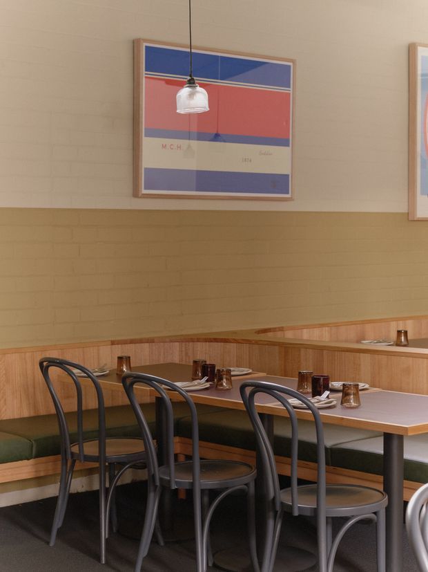 A warmly lit restaurant dining area with built-in banquette seating, timber wall panelling, painted brick walls, dark bentwood-style chairs, and simple set tables. Framed geometric artwork and pendant lighting add a refined mid-century-inspired character.