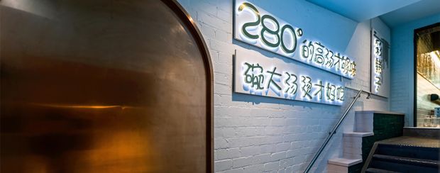 Interior view of a vividly lit commercial space with a blue-painted exposed brick wall, illuminated signage, a large curved bronze-toned architectural panel or doorway, and a narrow staircase with a metal handrail leading upward. The space has a dramatic, contemporary hospitality or retail character, with warm light reflecting from the metallic feature and daylight visible through glazing at right.