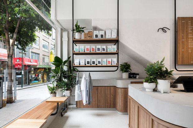 Bright contemporary commercial interior, likely a retail or hospitality counter space, with floor-to-ceiling street-facing glazing, white walls, light terrazzo-style flooring, curved timber cabinetry, a white counter, black metal shelving, and abundant potted greenery. Product packages and exterior street activity are visible but secondary to the fitted interior.