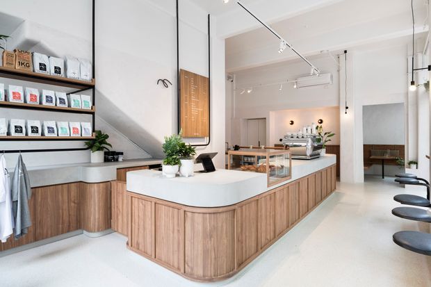 Bright contemporary café or specialty retail interior with a large rounded island counter, vertically grooved timber cabinetry, white benchtops, open wall shelving displaying packaged goods, and a row of black stools. White walls, exposed ceiling services, track lighting, glass display cases, and indoor plants create a clean, refined commercial fit-out.