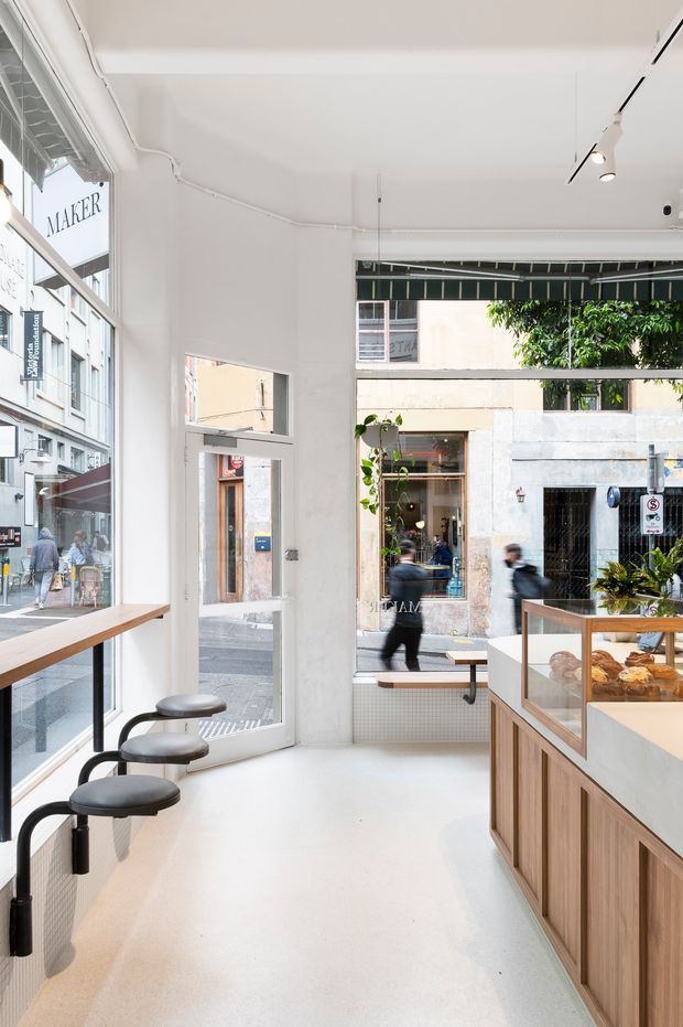 Bright, narrow contemporary café or bakery interior with full-height street-facing glazing, white walls and ceiling, pale seamless flooring, a timber wall-mounted counter with black stools, and a built-in timber service counter with a glass pastry display. The space opens directly onto an urban street and includes a linear track light and understated built-in joinery.