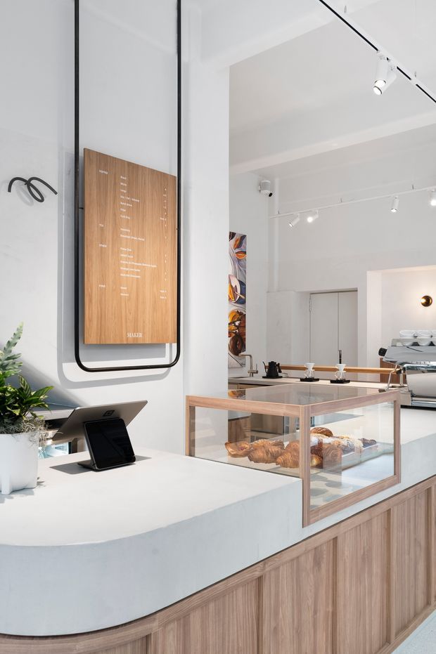 Bright contemporary bakery or café interior featuring a curved white service counter with wood-grain cabinetry, a glass pastry display case, espresso equipment, track lighting, and a minimalist wood menu board with black metal framing.