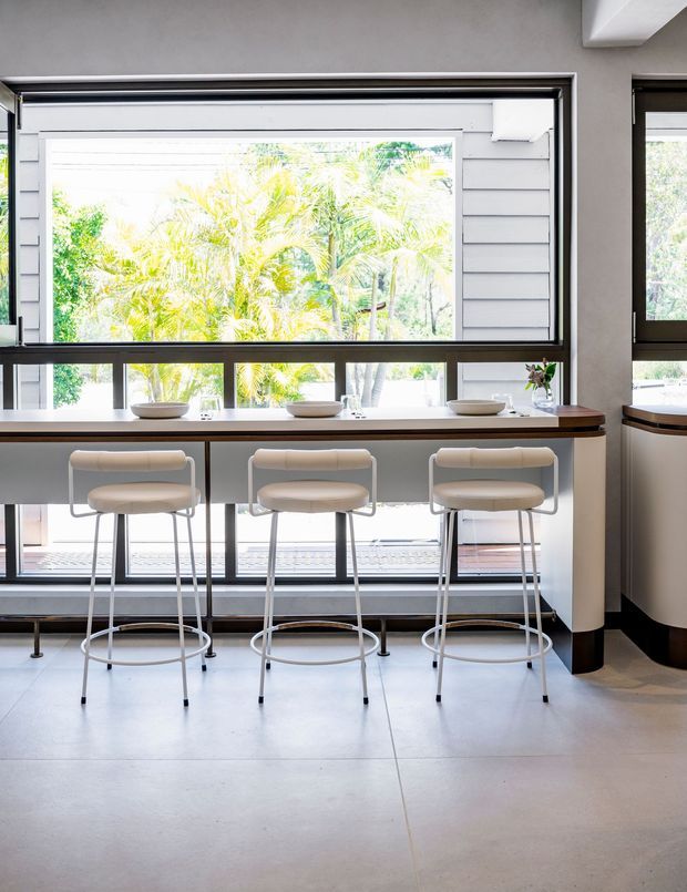 Bright contemporary kitchen or dining nook with a long timber-topped breakfast bar and three upholstered bar stools positioned in front of large windows. The space features pale smooth flooring, dark-framed glazing, light horizontal wall cladding, white walls, and abundant greenery visible outside.