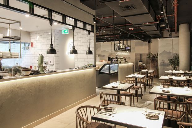 Contemporary industrial-style restaurant interior with an open kitchen and long illuminated textured counter, black pendant lights, exposed ceiling services, tiled walls, and a dining area furnished with timber chairs and white tables. Decorative screens and greenery add warmth to the otherwise neutral, utilitarian fit-out.