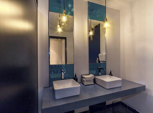A contemporary bathroom vanity with two white vessel basins set on a dark grey stone-look floating benchtop. Tall mirrors, blue glazed tile splashbacks, black tapware, and warm pendant lights create a refined, slightly dramatic interior.