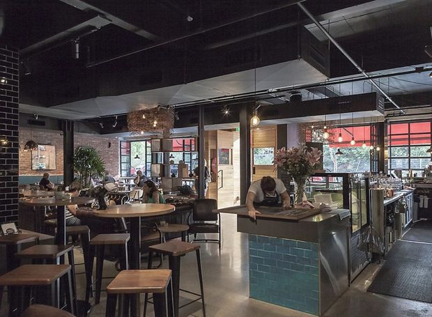 A dark, industrial-style café or restaurant interior with an open dining area and service counter. The space features exposed black ceilings and services, black-painted brick walls, large industrial windows, timber tables and stools, pendant lighting, indoor planting, and a green glazed-tile counter front.