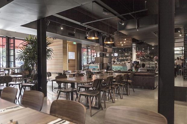 A spacious contemporary commercial dining and bar interior with numerous timber tables and chairs, a long bar, exposed black ceiling services, dark structural columns, pendant lighting, and a textured feature wall behind the bar. Large glazed openings bring in daylight, while indoor plants and warm timber finishes soften the industrial fit-out.